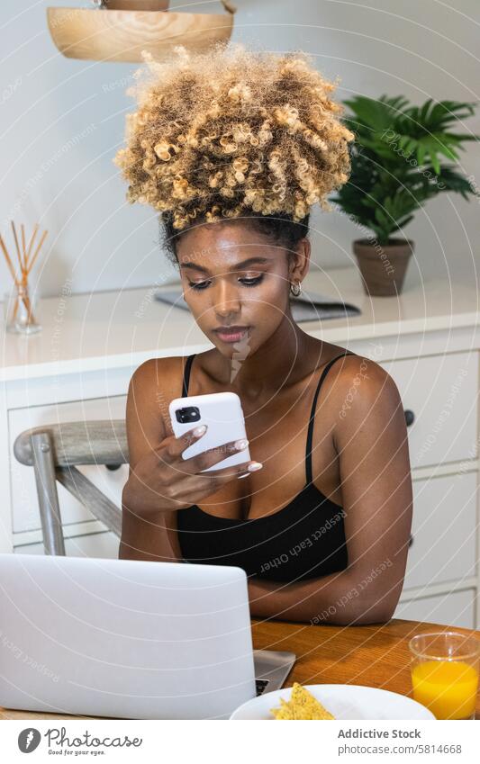 Schwarze seriöse Freiberuflerin mit Smartphone am Tisch beim Frühstück Frau benutzend freiberuflich zuschauen Laptop Morgen ernst Internet Saft Apparatur