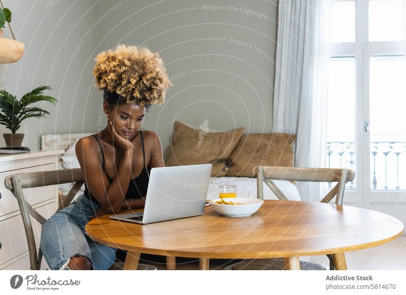 Schwarze Frau mit Laptop auf einem Stuhl sitzend beim Frühstück zuschauen Arbeit benutzend abgelegen online beschäftigt Morgen Gerät Browsen Apparatur heimwärts