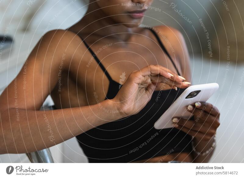 Anonyme schwarze Frau, die ein Smartphone benutzt, während sie auf einem Stuhl sitzt benutzend Internet online Textnachricht soziale Netzwerke zuschauen