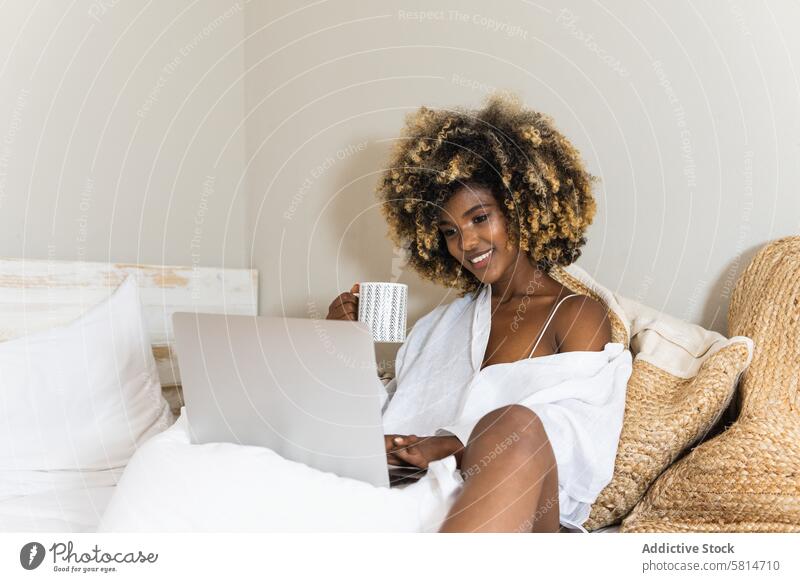 Schwarze Frau trinkt Kaffee, während sie mit einem Laptop auf dem Bett sitzt trinken benutzend Morgen Komfort Schlafzimmer Glück Kälte attraktiv Appartement