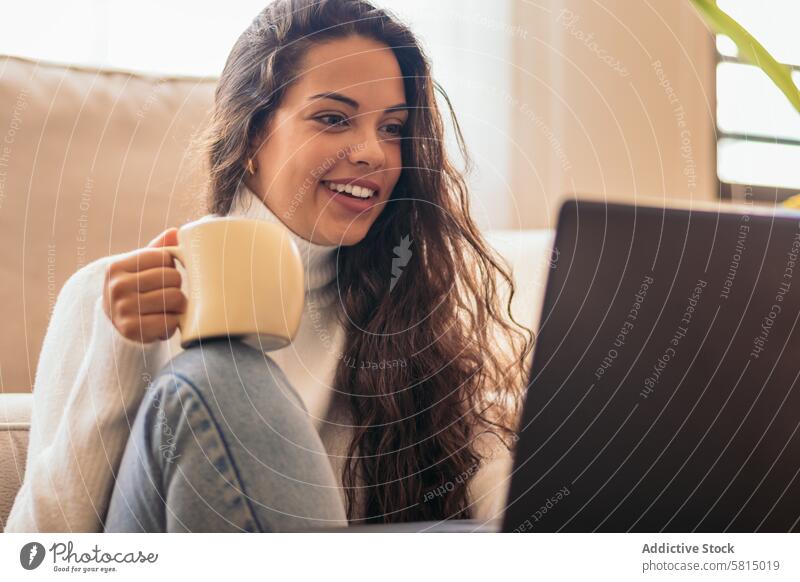 Junge Frau genießt eine Tasse Kaffee, während sie einen Laptop benutzt" Sofa heimwärts Technik & Technologie Produktivität abgelegen Anschluss Drahtlos modern