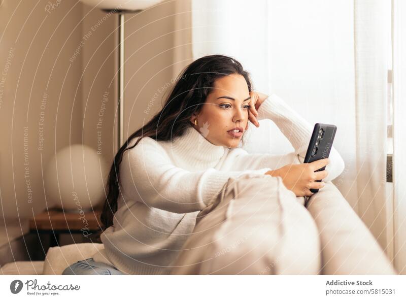 Vernetztes Zuhause: Junge Frau sitzt auf dem Sofa und benutzt ihr Smartphone Technik & Technologie Handy soziale Netzwerke digitaler Lebensstil