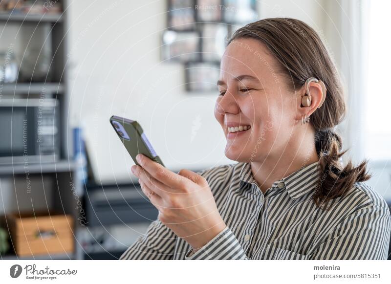 Frau mit Hörgerät und Smartphone Gehör Ohr Person benutzend taub Unterstützung Gesundheit Gerät Erwachsener Behinderung 30s weiß Lächeln Porträt Nachricht Hemd