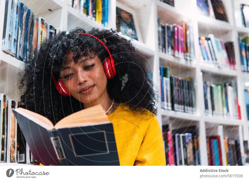 Zufriedene ethnische Studentin beim Lesen eines Lehrbuchs in der Bibliothek Frau lesen Buch Schüler Lächeln Bildung Glück Leseratte Bücherschrank positiv trendy