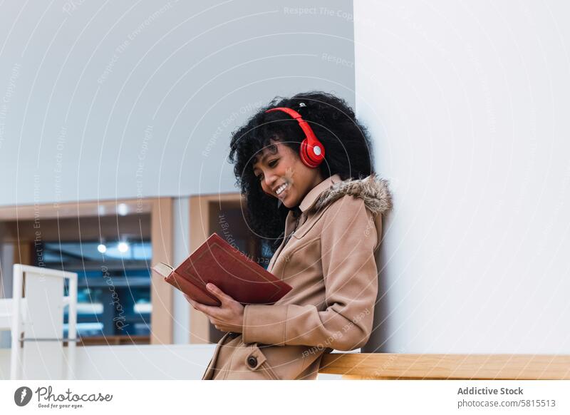 Zufriedene ethnische Studentin beim Lesen eines Lehrbuchs in der Bibliothek Frau melomaniac Regal Schüler Buch Bücherschrank Kopfhörer positiv wählen