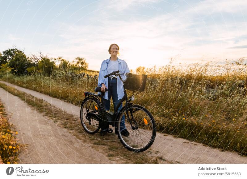blonde Frau stehend mit einem Fahrrad in einem Feld bei Sonnenuntergang jung Mädchen Mode Sommer Stehen Natur hübsch Landschaft Lifestyle reisen schön Glück