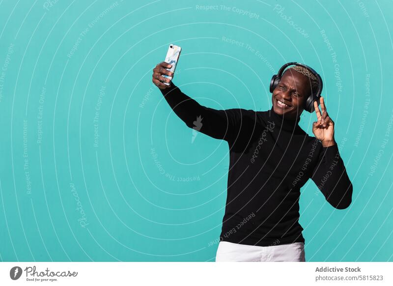 Fröhlicher junger schwarzer Mann, der mit Kopfhörern Musik hört und ein Selfie auf der Straße macht zwei Finger Lächeln zuhören Smartphone Freude heiter trendy