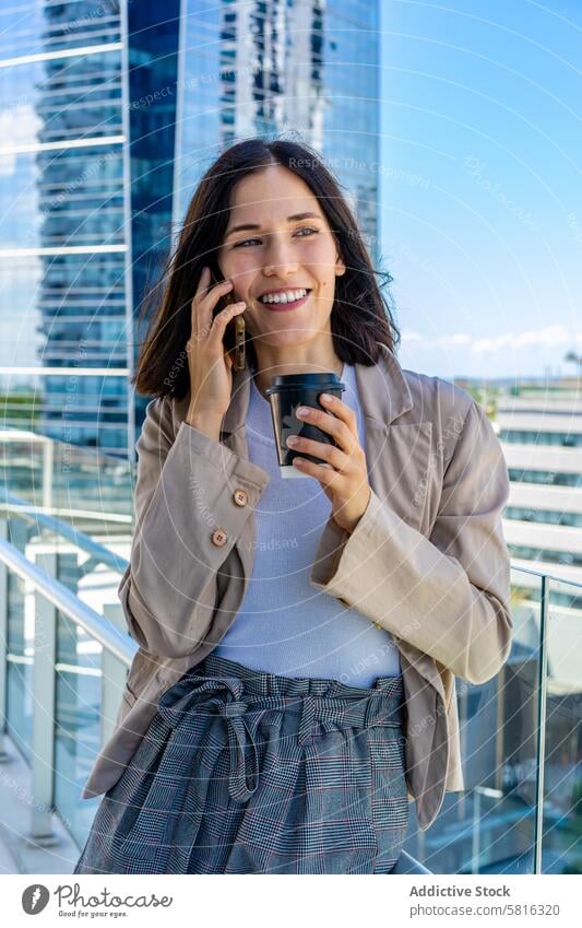 Eine Frau steht auf einem Balkon und telefoniert in ihrer Büropause mit ihrem Handy.  In der Hand hält sie einen Pappbecher mit Kaffee. modern attraktiv