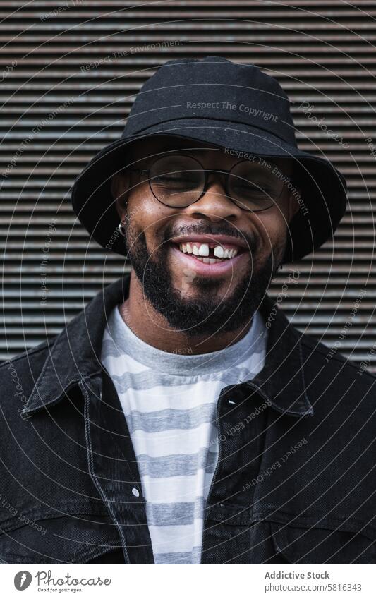 Lächelnder schwarzer Hipster-Mann mit Hut schaut in die Kamera Glück trendy heiter Vollbart Brille Optimist männlich Afroamerikaner ethnisch Outfit Stil positiv
