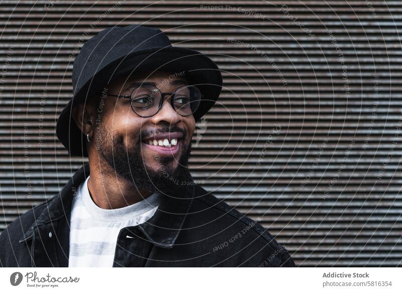 Lächelnder schwarzer Hipster-Mann mit Hut schaut weg Glück trendy heiter Vollbart Brille Optimist männlich Afroamerikaner ethnisch Outfit Stil positiv Freude