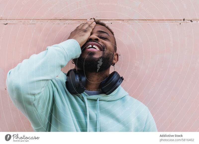 Erfreuter ethnischer Hipster-Mann bedeckt Auge heiter gestikulieren Abdecköse Glück Kopfhörer Vollbart lässig Erwachsener Afroamerikaner schwarz Spaß haben