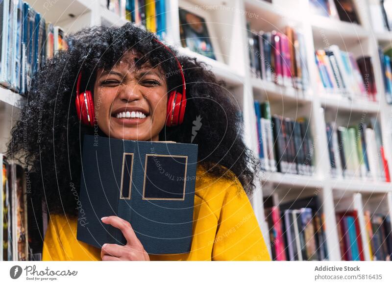 Zufriedene ethnische Studentin mit Buch in der Bibliothek Frau zuhören lesen Musik Schüler Lächeln Bildung Glück Leseratte Bücherschrank positiv trendy jung