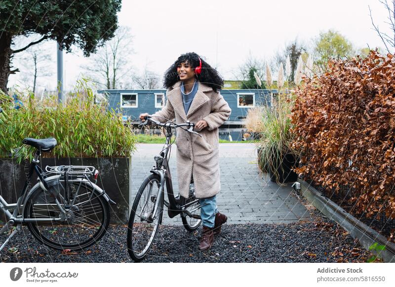 Fröhliche schwarze Frau mit Kopfhörern und Fahrrad benutzend Musik zuhören Kälte Großstadt afroamerikanische Frau Straße Lächeln Spaziergang schlendern positiv