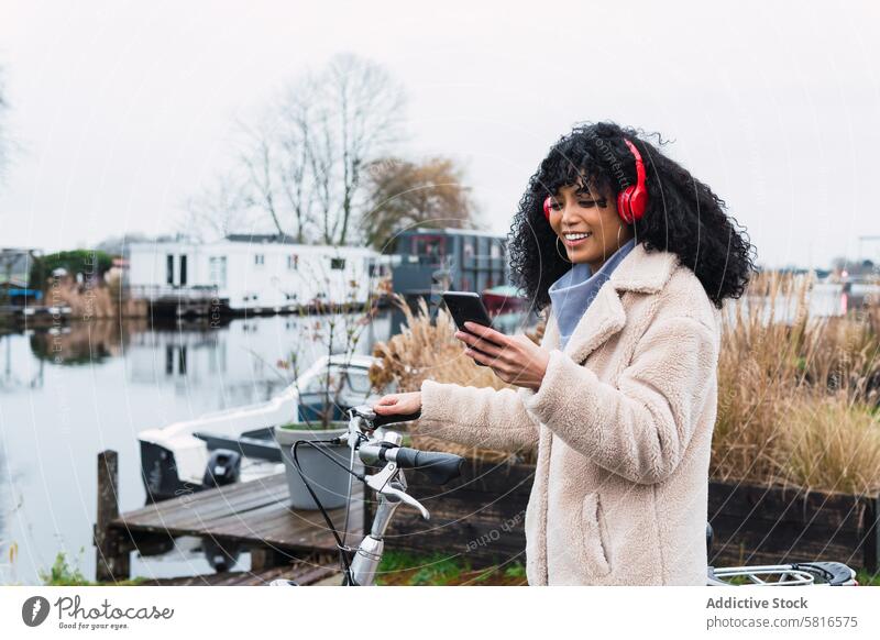 Vergnügte schwarze Frau mit Smartphone auf dem Fahrrad Kopfhörer benutzend Musik zuhören Spaziergang Mitfahrgelegenheit Kälte afroamerikanische Frau Großstadt
