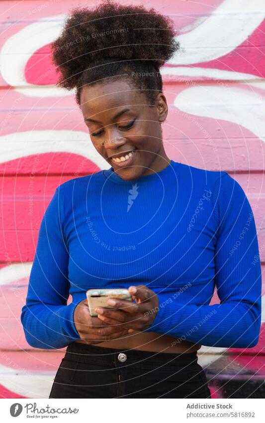 Zufriedene schwarze Frau, die in der Stadt auf ihrem Smartphone surft Browsen zuschauend Graffiti Wand Großstadt Straßenkunst heiter ethnisch Afroamerikaner