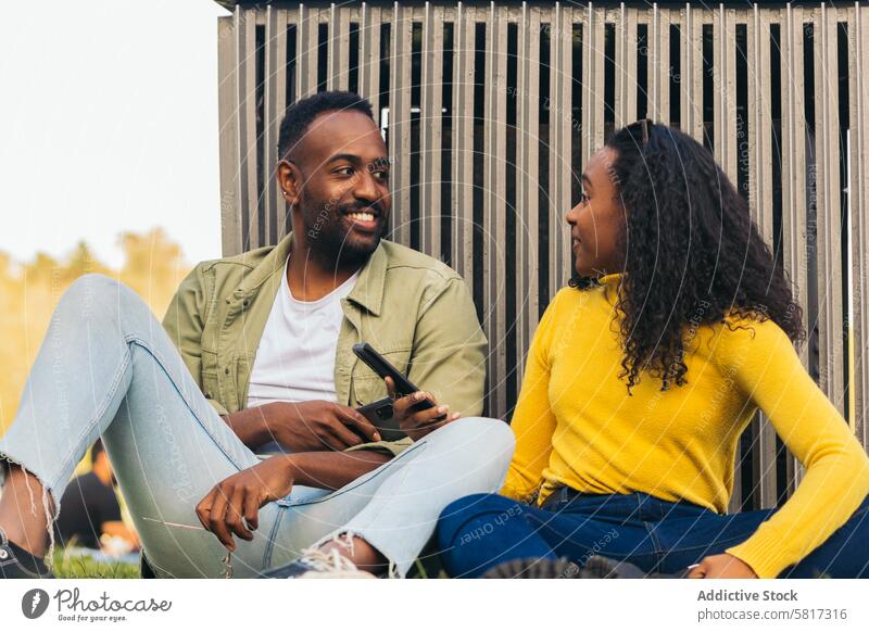 Schwarzes Paar sitzt in einem Park und benutzt ein Handy jung Mobile Telefon Freunde Sitzen Internet online Mann Frau Smartphone im Freien Afrikanisch Freizeit