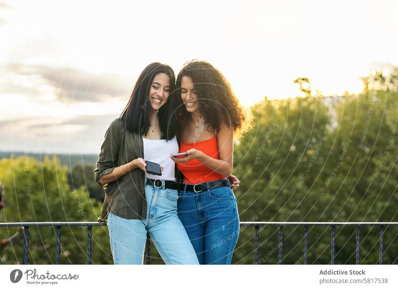 zwei junge Frauen lächelnd und mit ihren Handys auf einer Terrasse. mit dem Sonnenuntergang Menschen Smartphone Technik & Technologie Mobile benutzend