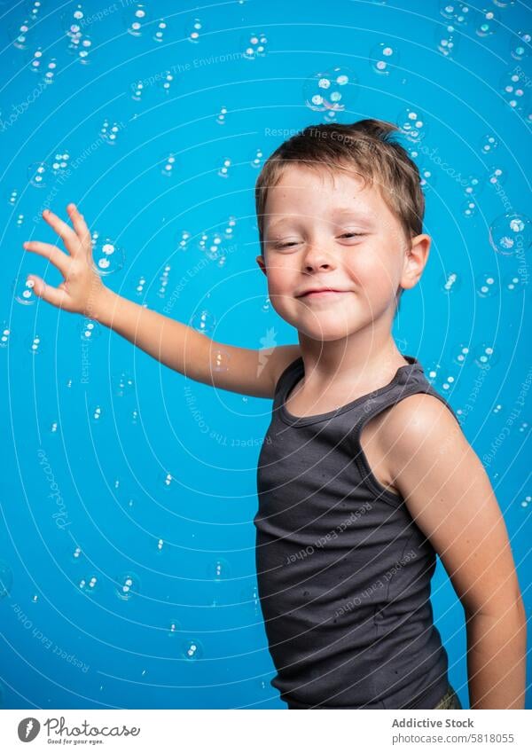 Fröhlicher Junge spielt mit Seifenblasen auf blauem Hintergrund Kind Blasen spielen Studioaufnahme Blauer Hintergrund Inhalt spielerisch Tanktop erreichend