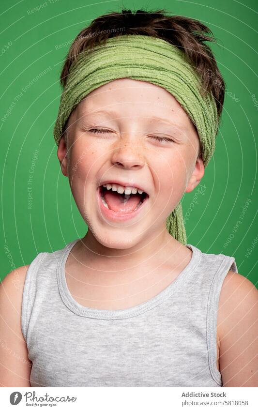 Fröhlicher Junge, der mit geschlossenen Augen im Studio lacht Kind Lachen heiter Glück Freude Studioaufnahme grüner Hintergrund Stirnband jung männlich Spaß