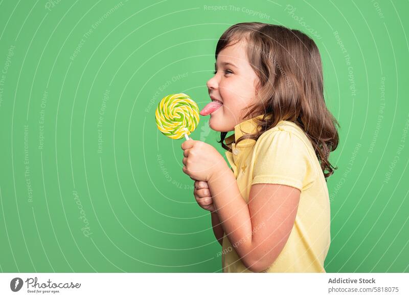 Fröhliches Mädchen genießt einen Swirl-Lollipop im Studio Kind Verwirbelung heiter Studioaufnahme grüner Hintergrund gelbes Hemd Genuss Bonbon süß Snack lecken