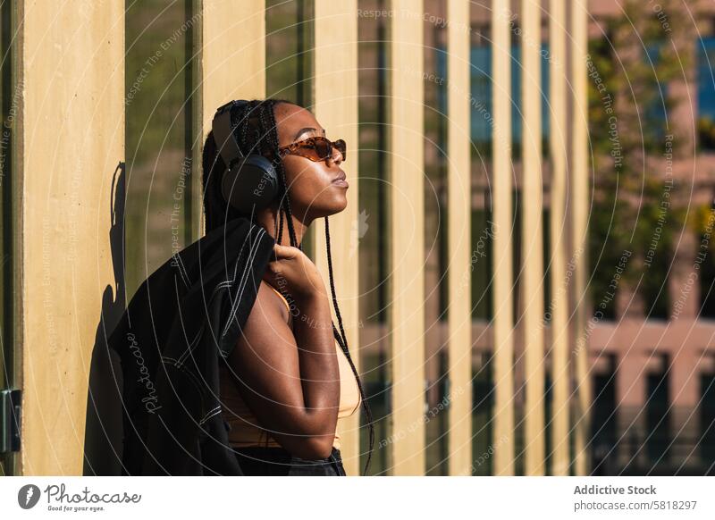 Ethnische Frau hört Musik vor einem modernen Gebäude ruhen Straße zuhören fettarm Wand jung Großstadt urban trendy Klang Audio Kopfhörer Zeitgenosse Jacke