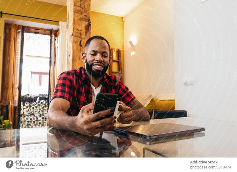 Afroamerikanischer Mann, der sein Smartphone benutzt, während er zu Hause Kaffee trinkt Laptop Telearbeit Computer Afroamerikaner heimwärts Internet