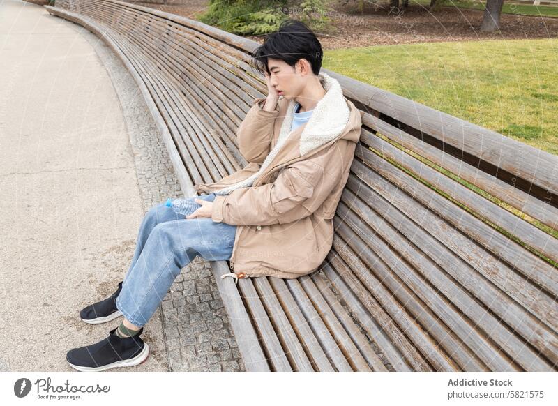 Junger Mann fühlt sich auf einer Parkbank unwohl, Thema Hypoglykämie jung asiatisch Bank Sitzen Unbehagen Kopf Hand Wasserflasche Jeanshose Jacke im Freien Tag