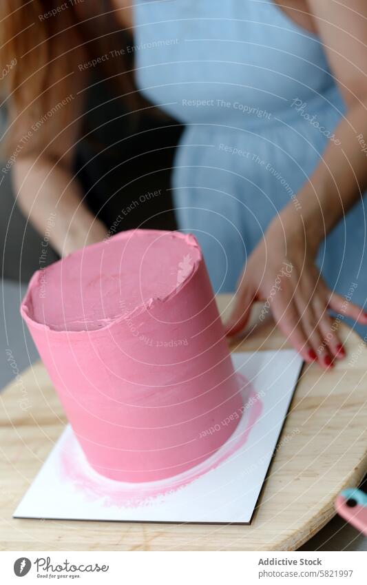 Dekorateurin glasiert einen rosa Kuchen auf einem Holzbrett Zuckerguß hölzern Holzplatte Konditorei Schürze blau Kunst backen dekorierend Dessert Gebäck