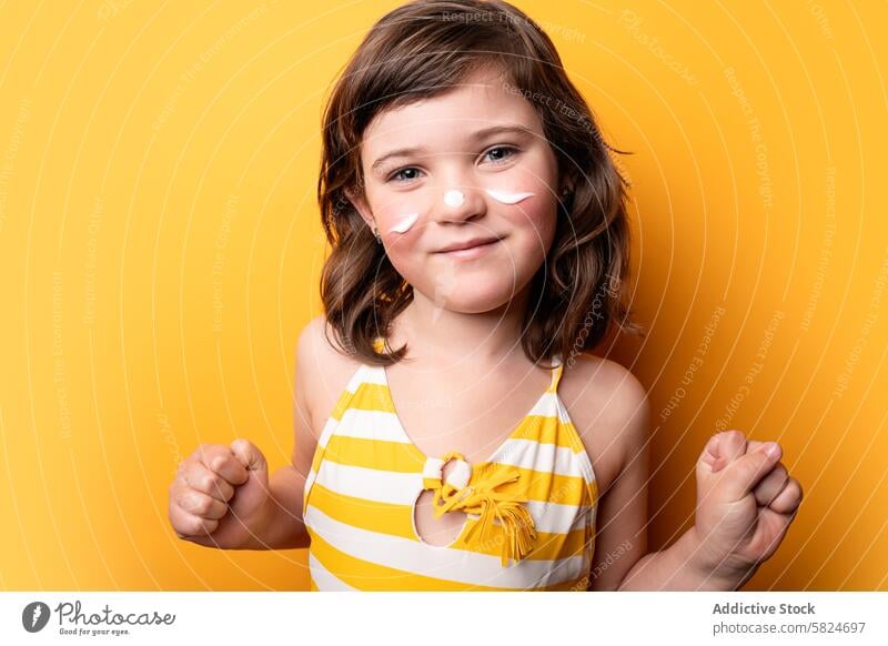 Junges Mädchen mit Sonnencremestreifen auf gelbem Hintergrund Kind heiter posierend Sommer Schutz Hautpflege Glück freudig pulsierend Badeanzug gestreift