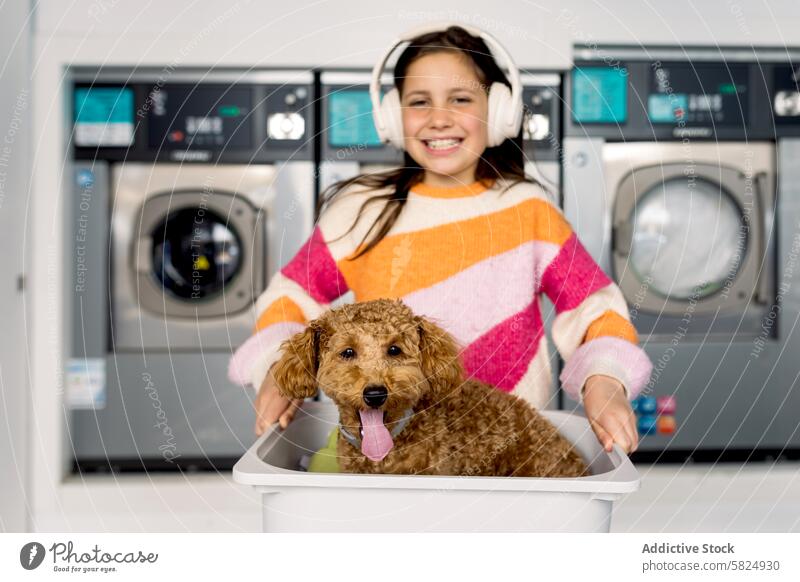 Preteen Mädchen und Hund haben Spaß in der Waschküche Kind Lächeln Korb Kopfhörer spielerisch in die Kamera schauen im Innenbereich Waschmaschinen