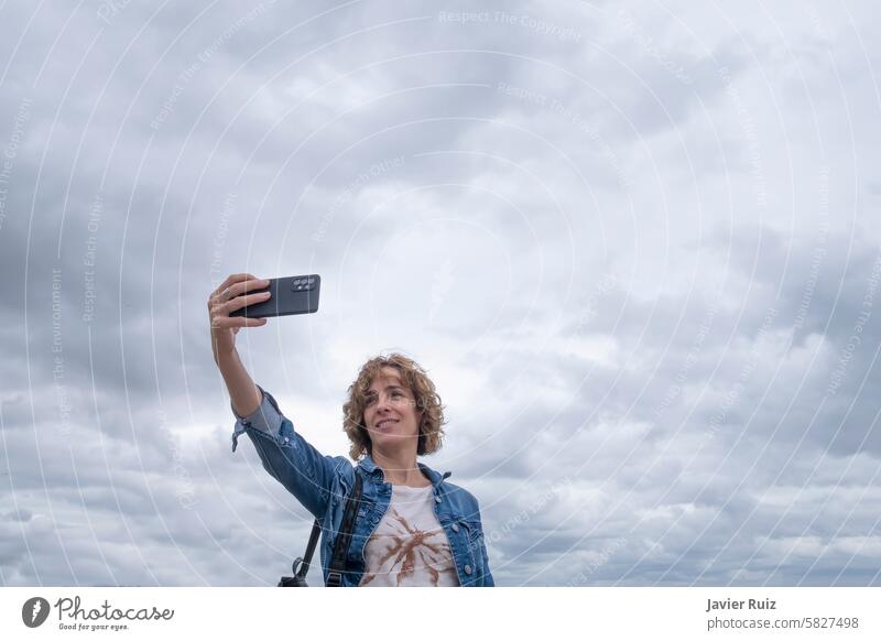 eine lächelnde Frau mittleren Alters in legerer Kleidung in den Fünfzigern macht ein Selfie mit ihrem Mobiltelefon vor einem bewölkten Himmel, Kopie des Raums