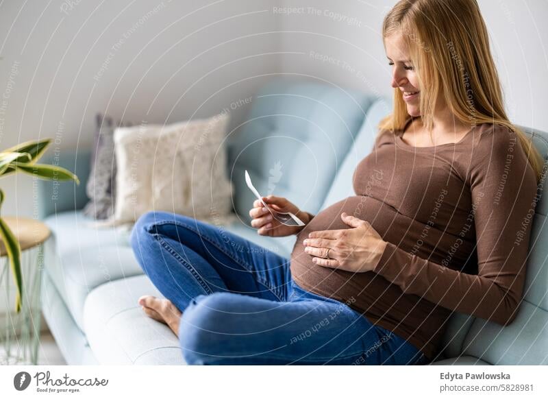 Schwangere Frau, die zu Hause auf dem Sofa sitzt und sich das Ultraschallbild ihres Babys ansieht Schwangerschaft schwanger Erwachsener Vorfreude wartend Bauch