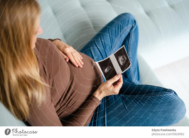 Schwangere Frau, die zu Hause auf dem Sofa sitzt und sich das Ultraschallbild ihres Babys ansieht Schwangerschaft schwanger Erwachsener Vorfreude wartend Bauch