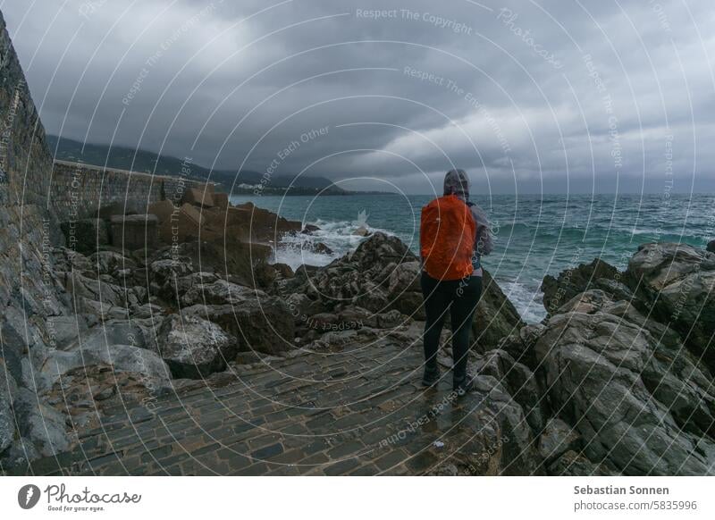 Tourist steht an der Küste in der Nähe der mittelalterlichen Hafenmauer am Mittelmeer an einem bewölkten Regentag, Cefalu, Sizilien, Italien MEER Wand reisen