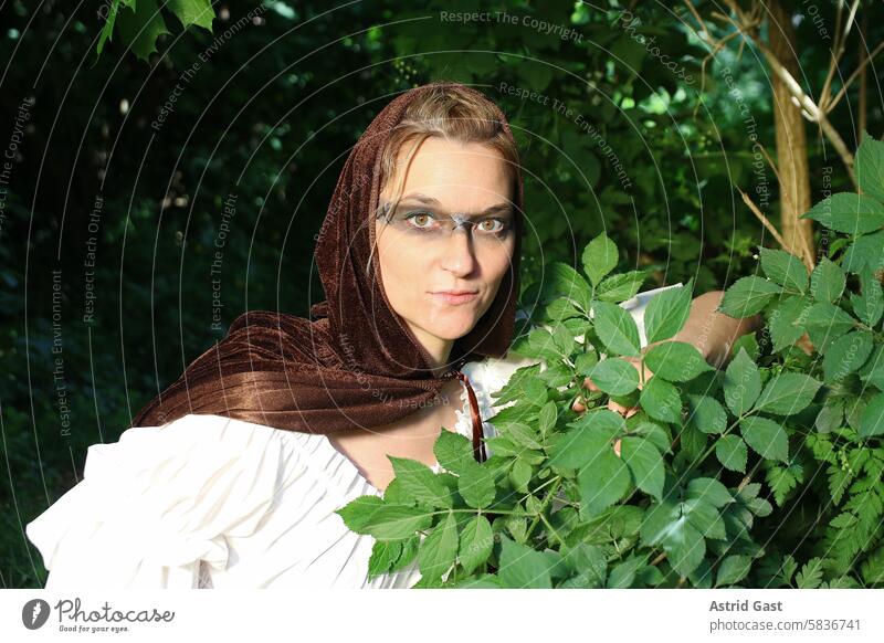 Eine Frau, als mittelalterliche Jägerin verkleidet, schaut aus dem Wald frau hexe elfe zauberin magierin gesicht umhang elfenhaft hexenhaft fantasie jägerin