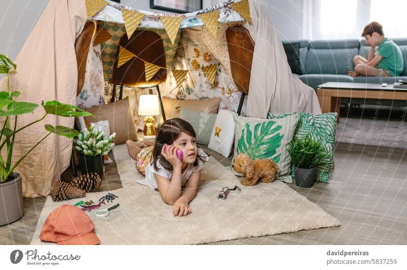 Kleines Mädchen mit Walkie-Talkie auf einem Teppich in einem Schutzzelt liegend, während es sich mit einem Jungen auf dem Sofa unterhält sprechend Spielen