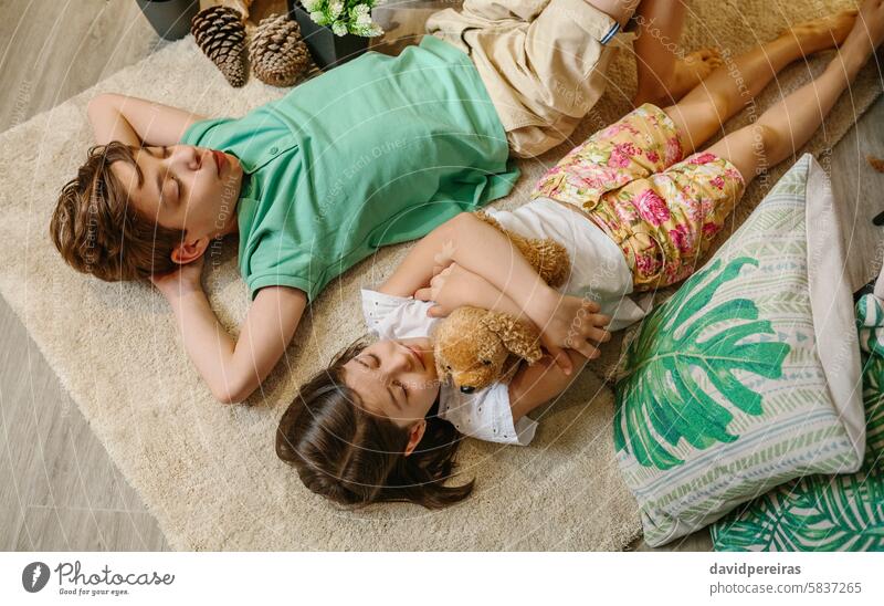 Draufsicht auf zwei Kinder mit geschlossenen Augen, die sich auf einem Teppich in einem gemütlichen Raum mit warmem Ambiente entspannen. Erholung Lügen Vorleger