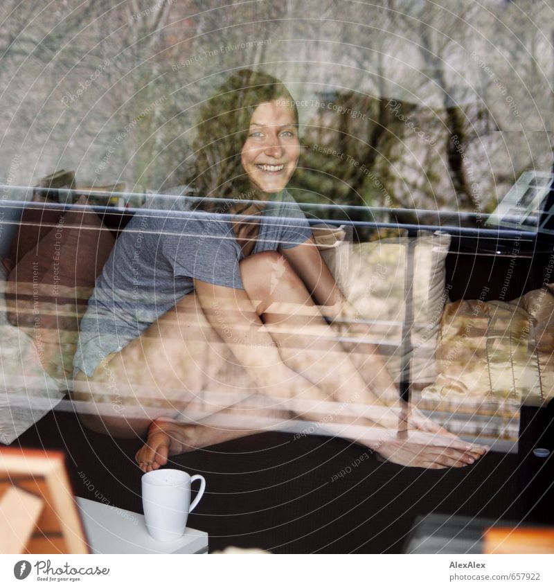 junge Frau hinter einer Fensterscheibe sitzt langbeinig und barfuß auf dem Sofa Junge Frau Jugendliche Körper Beine 18-30 Jahre Erwachsene T-Shirt Barfuß