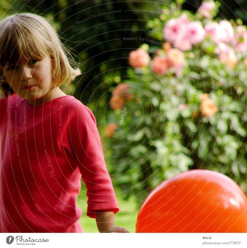 Blonde, süßes Mädchen steht mit rotem Luftballon in der Hand im Garten vor rosa Blumen in voller Blüte. Geburtstagskind draussen in der Natur freut sich über geschenkten Luftballon zu ihrem Fest.
