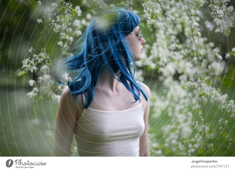 Frau mit blauen Haaren steht im Profil unter einem Baum mit weißen Blüten, sie schenkt ihren Kopf nach rechts und die Haare fliege mit Perücke Sommer Sonne