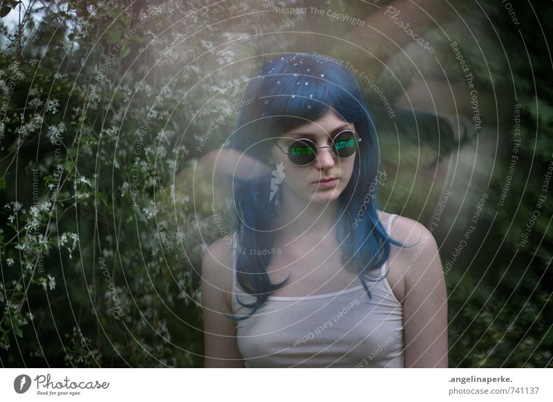 Frau mit blauen Haaren vor einem Busch mit weißen Blüten, sie trägt eine Sonnenbrille mit der Aufschrift "love" Perücke 70s Love psychedelisch Sommer