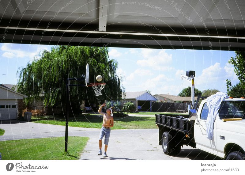 Junge mit freiem Oberkörper und Stirnband spielt vor der Garage Basketball. Neben ihm steht ein Pickup Truck Basketballkorb Amerika Korb USA Gras Garagentor